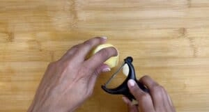 Hands peeling a potato with a black peeler on a wooden cutting board.